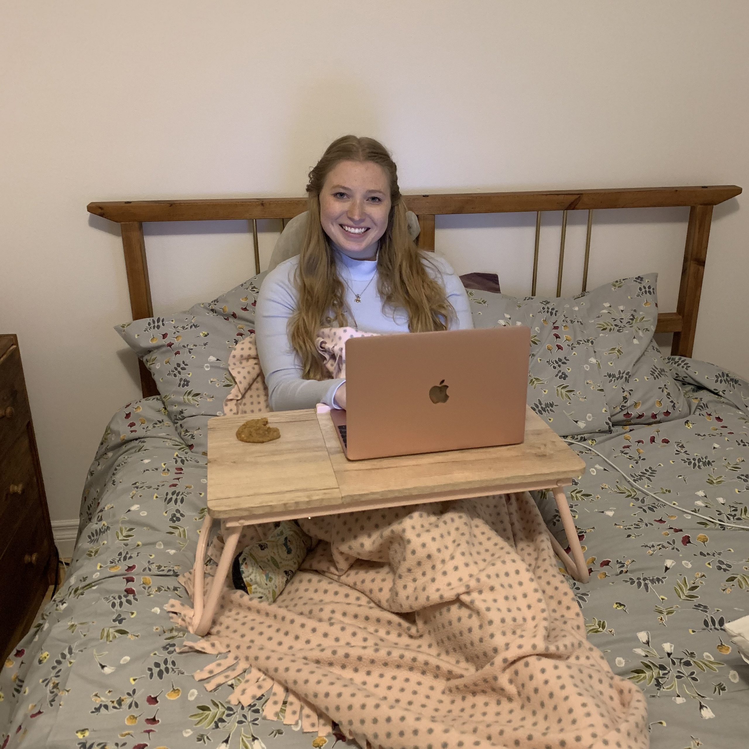 Kelcie on her bed and working Kelcie sitting up on her bed. She looks comfortable and wrapped in blankets. There is a bed tray propped in front of her with her laptop on it. The laptop is open and you can see the light from the screen hitting her face. She is smiling and looking directly at the camera.