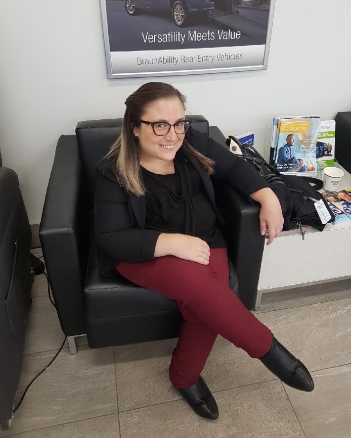 IMG_6516 Gabby sitting in a low black lounge chair with her legs crossed. She is looking at the camera and smiling. She has her hair pinned to the back, she is wearing glasses, a black cardigan, maroon pants and black boots.