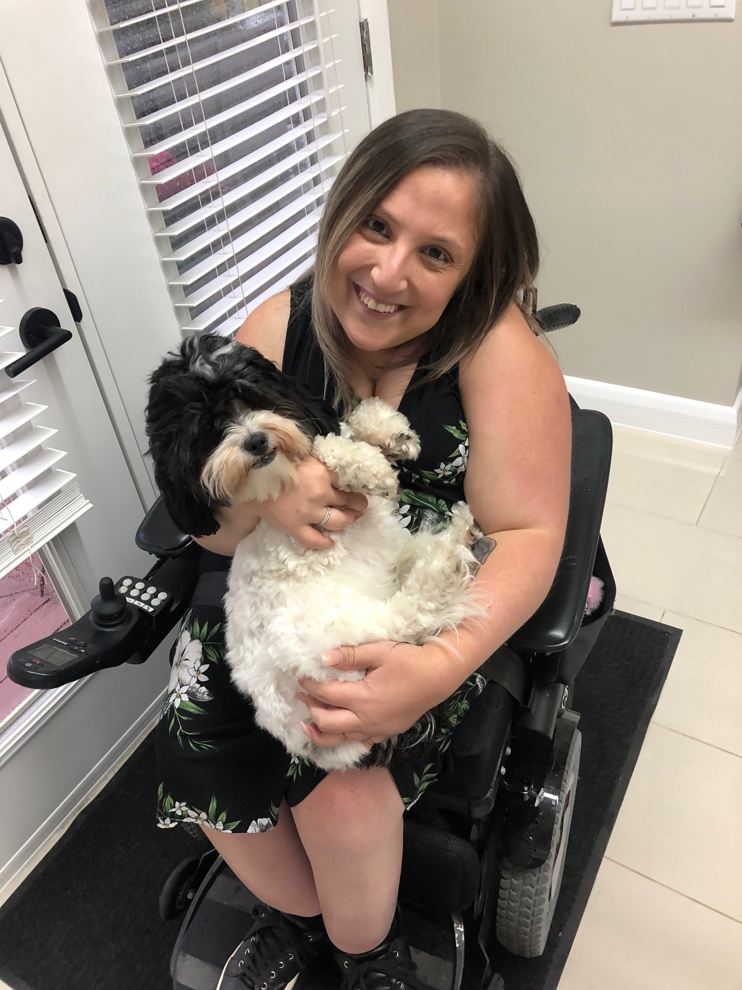 gabby Gabby in her wheelchair. She is looking at the camera with a sweet smile on her face while she is holding a dog on her lap.