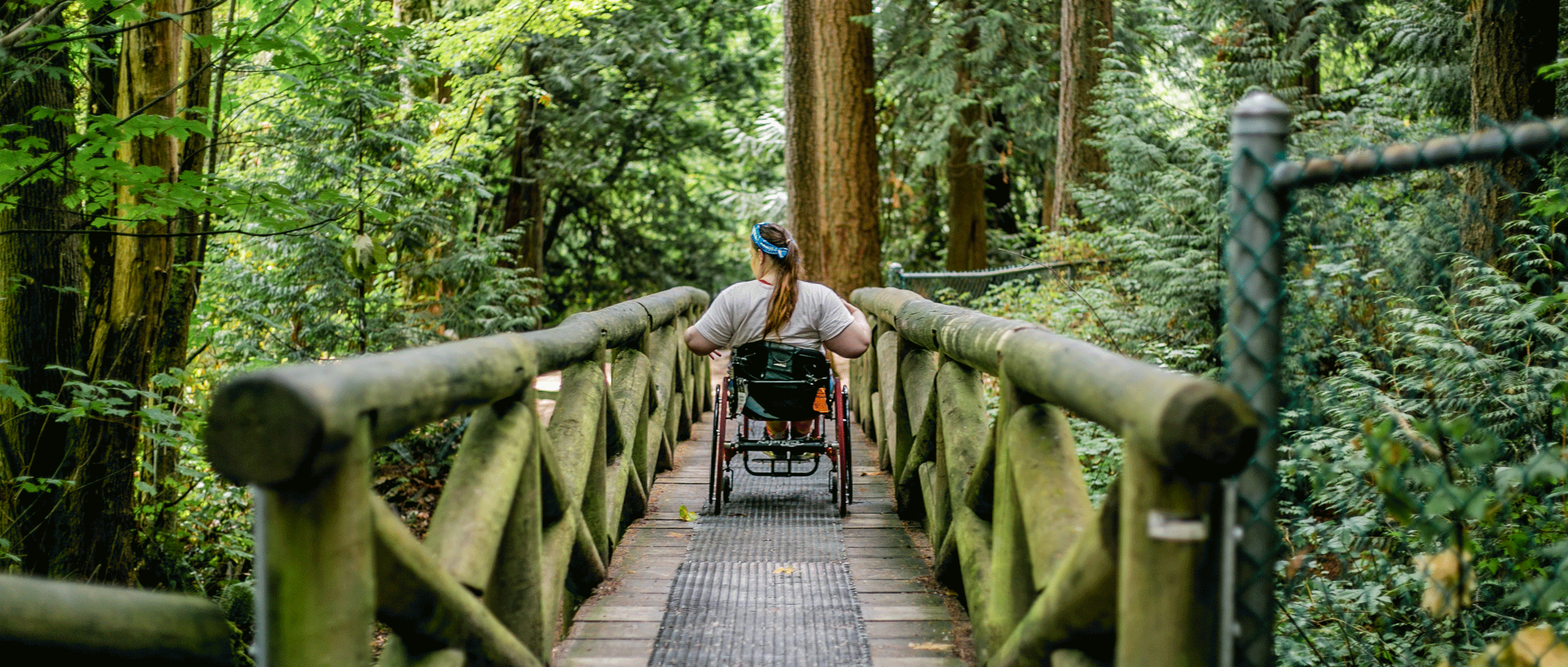 website-TRAILS-2021-SURREY---Photo-by-Alexa-Fernando---20 woman in manual wheelchair rolls through a mossy bridge in a forest