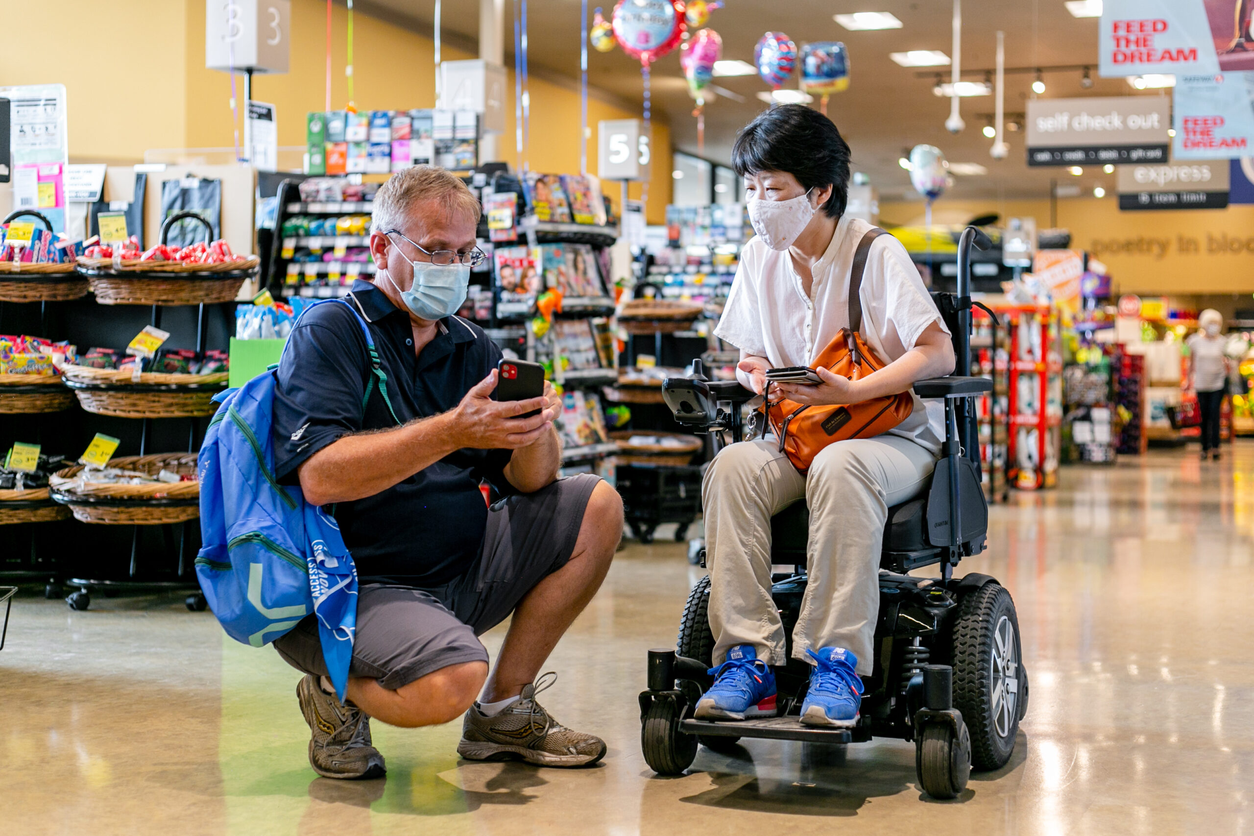 MOCA 2021 YYC - Photo by Alexa Fernando - 99 an asian woman sitting in a wheelchair and a white man both look at a mobile phone while inside a grocery store
