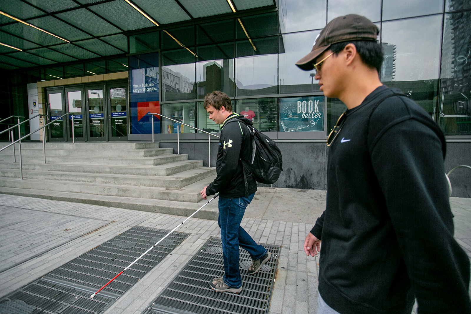 MOCA-2021-YYC-web---Photo-by-Alexa-Fernando---10 a blind man navigates a city sidewalk with his white cane alongside his non-disabled colleague. In the background is a building with stairs to enter.