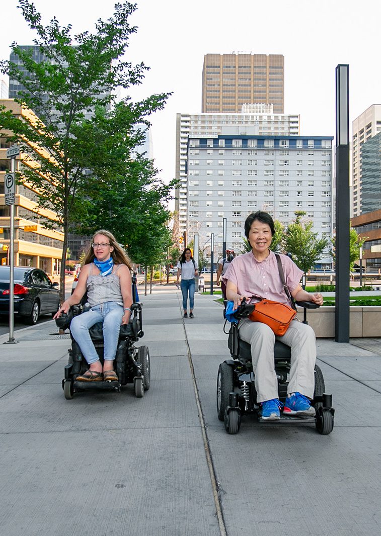 MOCA 2021 YYC web - Photo by Alexa Fernando - 36 two power wheelchair users drive toward the camera smiling on a city sidewalk